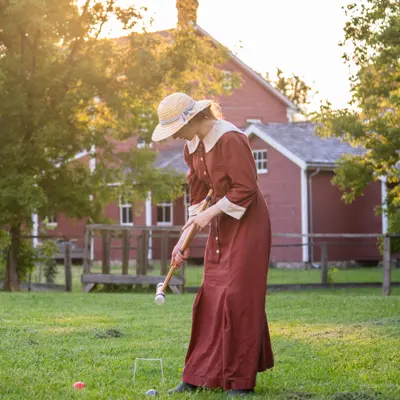 young woman in burgandy period dress and straw hat playing croquet